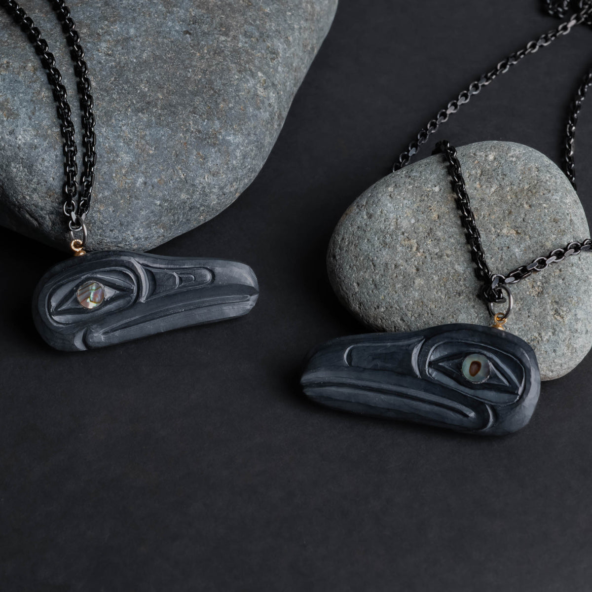 Two carved black stone pendants with abalone eye and black chain set against grey rocks on a black back ground.