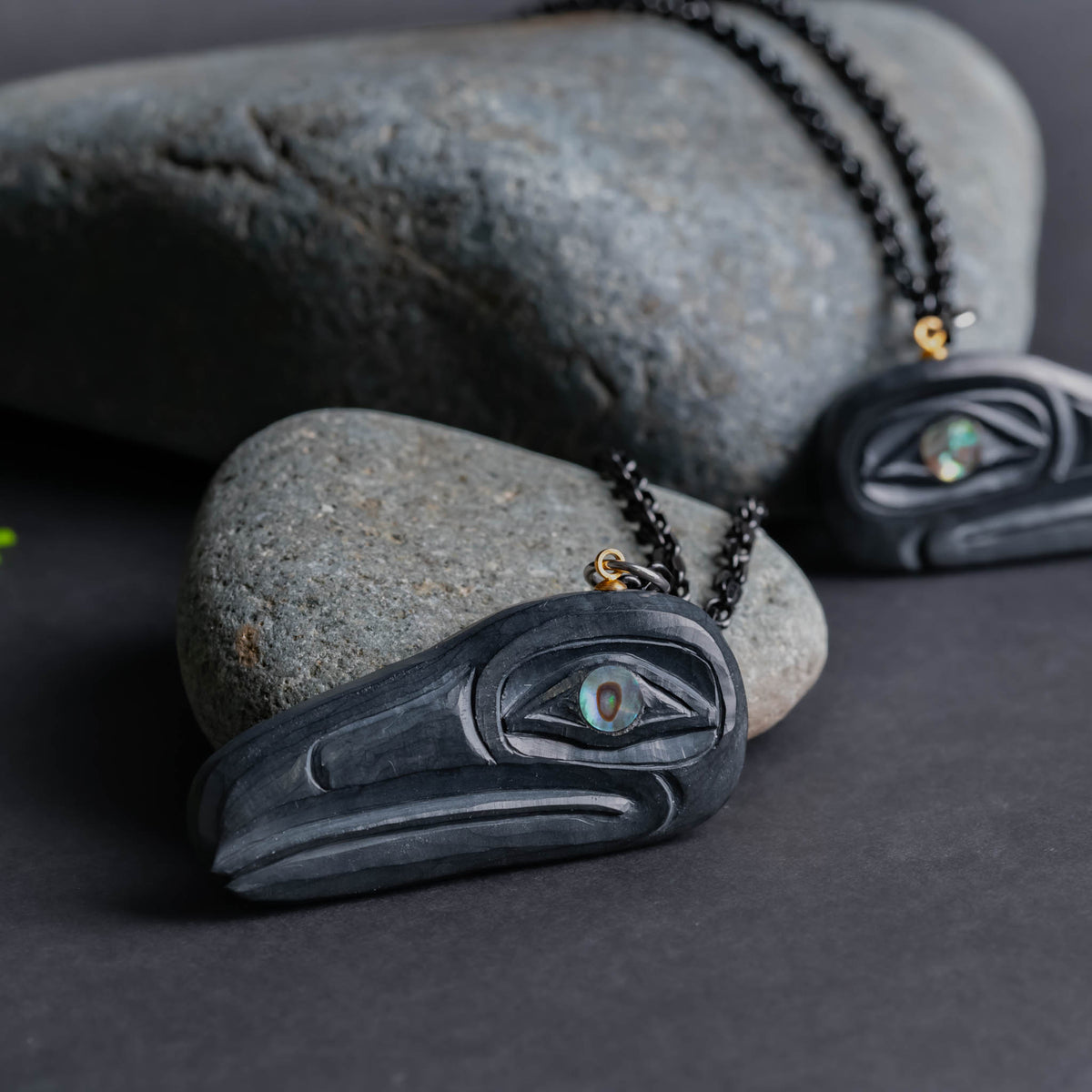 Haida Argillite Carving Raven Pendants with abalone eyes by Sean Brennan set against grey rocks on a black background.