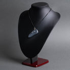 A carved stone raven necklace displayed on a black jewelry bust with a dark red base, photographed against a smooth gray studio background.