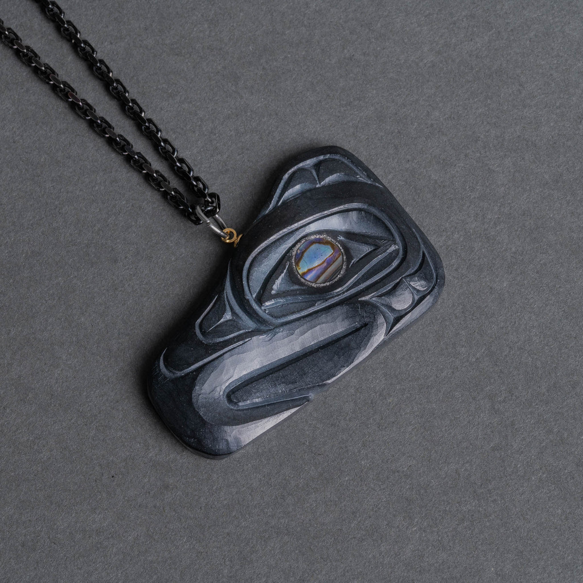 Haida Argillite Carving Thunderbird Pendant by Sean Brennan with abalone eye, hanging from a black chain on a dark grey background.