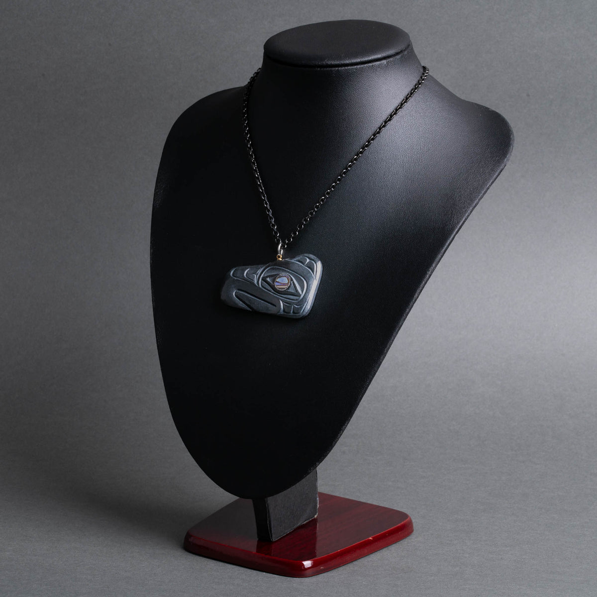 A dark gray carved thunderbird necklace displayed on a black jewelry bust with a red base, photographed against a neutral gray studio background.