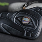 Close up of black stone carving with an iridescent a abalone eye with grey and green objects in the background.