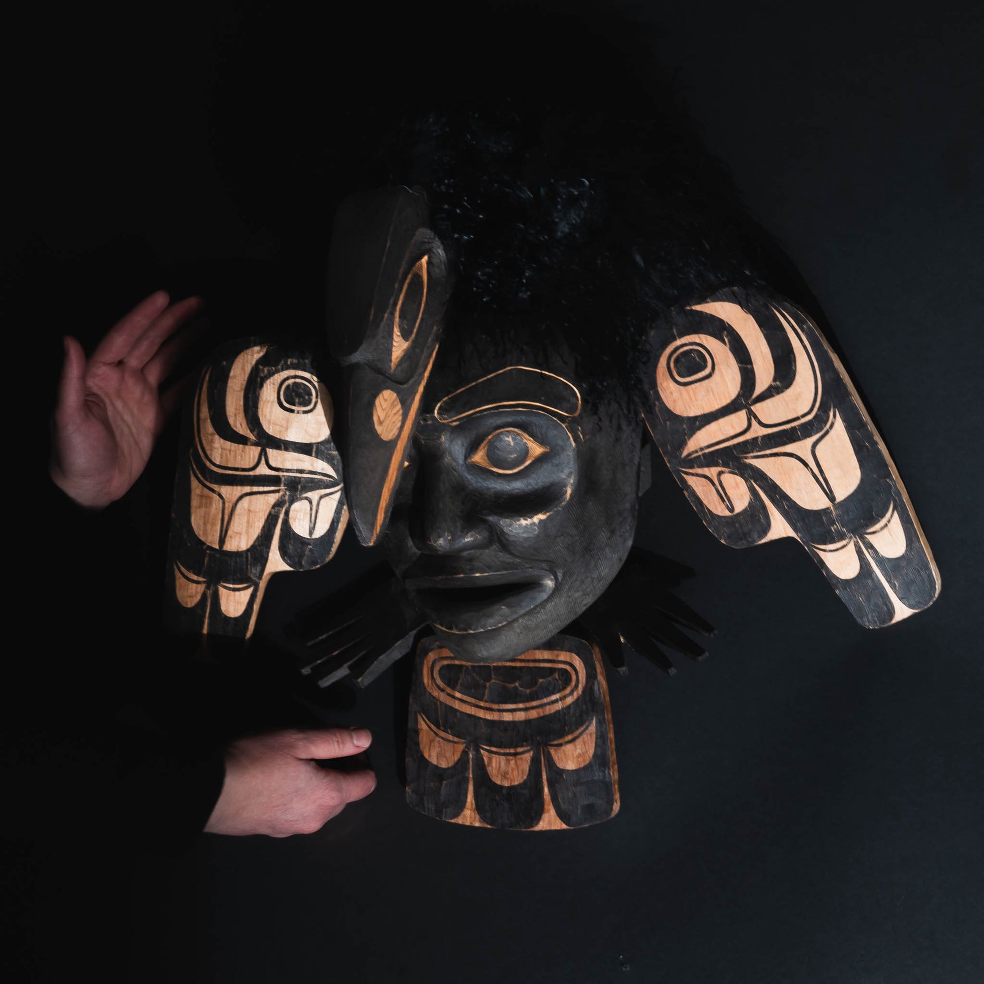 A black mask with wings and a human face being help by two hands.