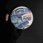 Person holding round painted panel with a figure holding a basket in blue, purple, and, orange.