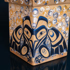 A close-up of a decorative square box with a painted Indigenous-style bird face and circular patterns in navy, gold, white, and tan on a dark reflective surface.