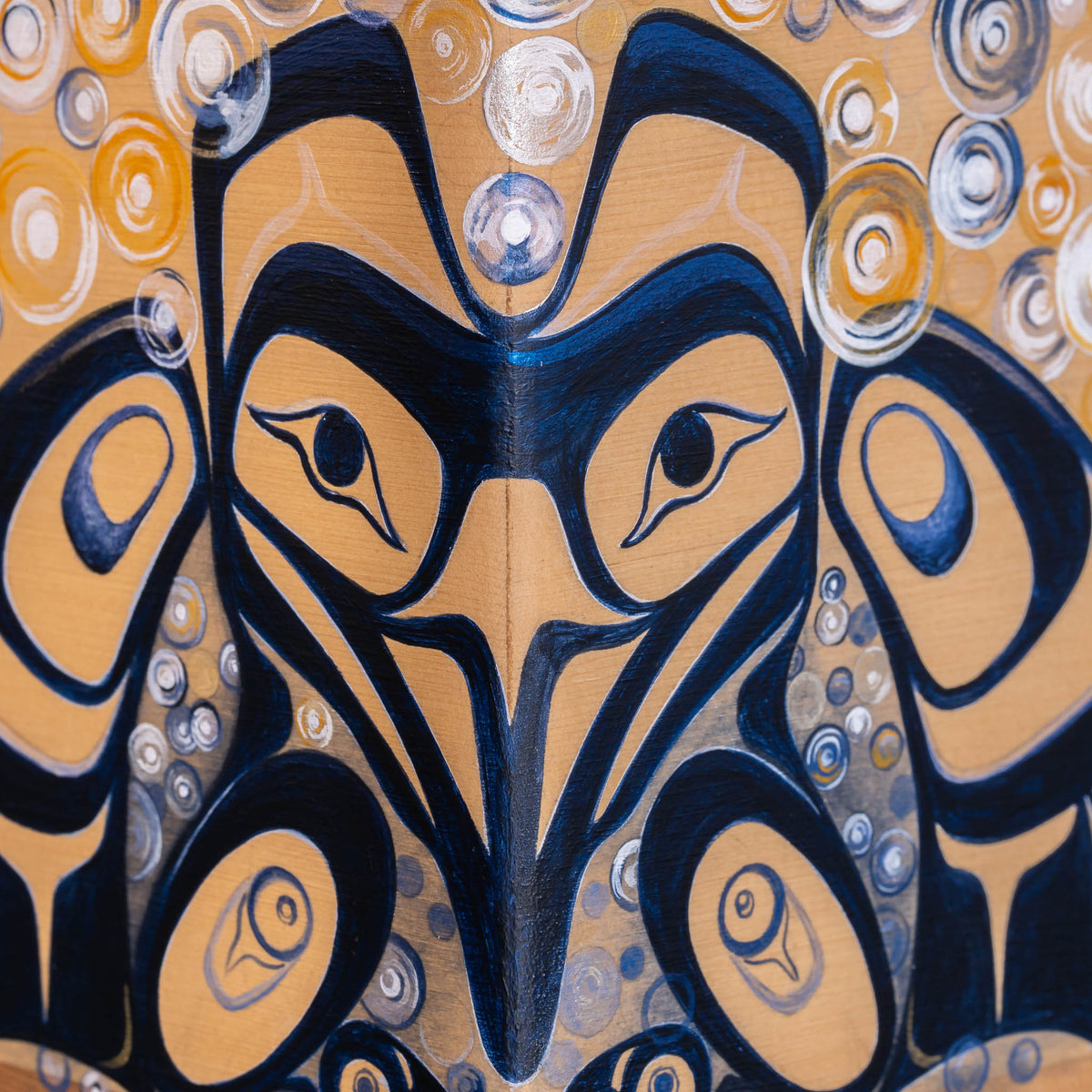 A close-up of a painted artwork featuring a stylized raven in dark navy blue and warm tan, surrounded by circular abstract patterns in tan, navy, white, and yellow.