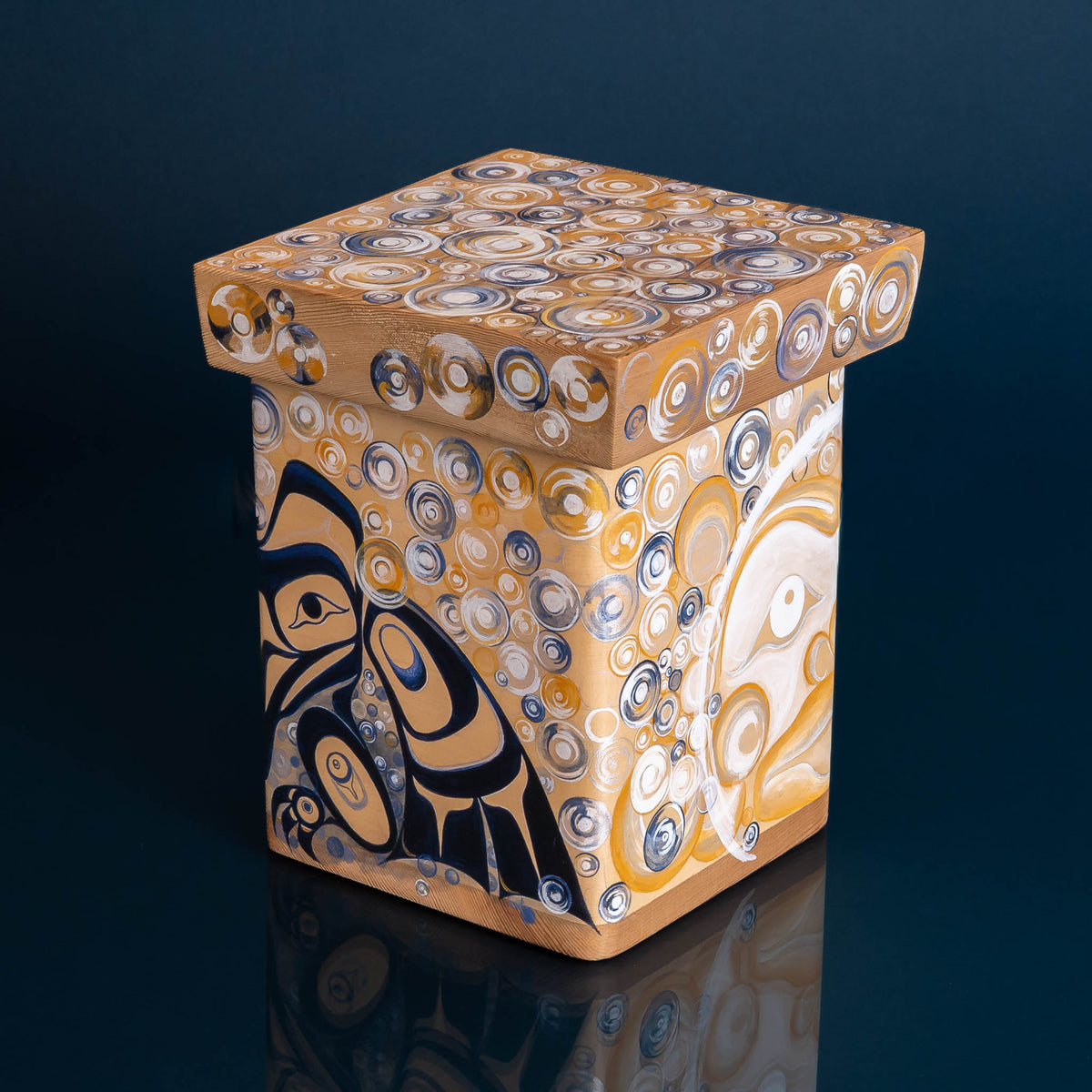 A wooden box painted in blues, yellows,ows, and whites, featuring circular patterns and two separate faces, on a dark blue background. 