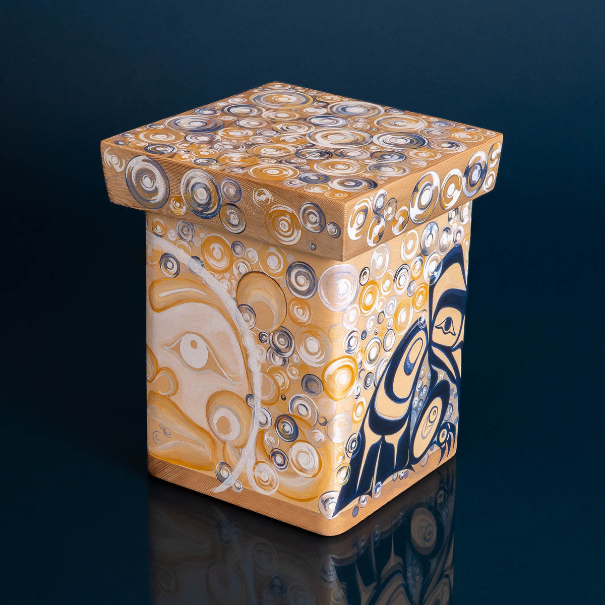 A wooden box painted showing two faces, one golden and one dark, with painted spheres around them on a dark blue background. 