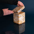 A handcrafted square wooden box with a lid, decorated with a stylized face and circular patterns in gold, beige, and silver, shown with hands lifting the lid against a dark blue background.