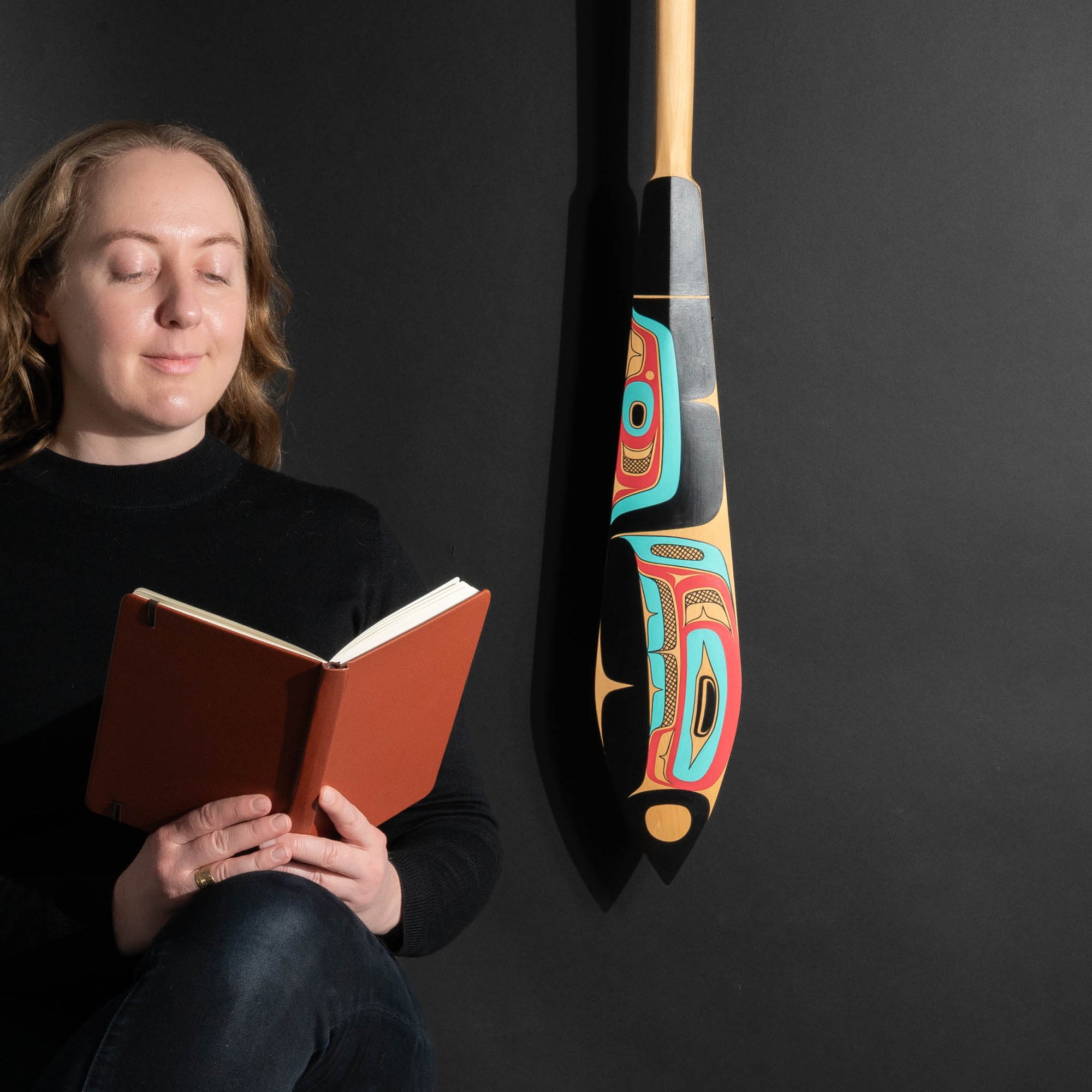 Person reading a book next to a colorful haida formline paddle on a black background