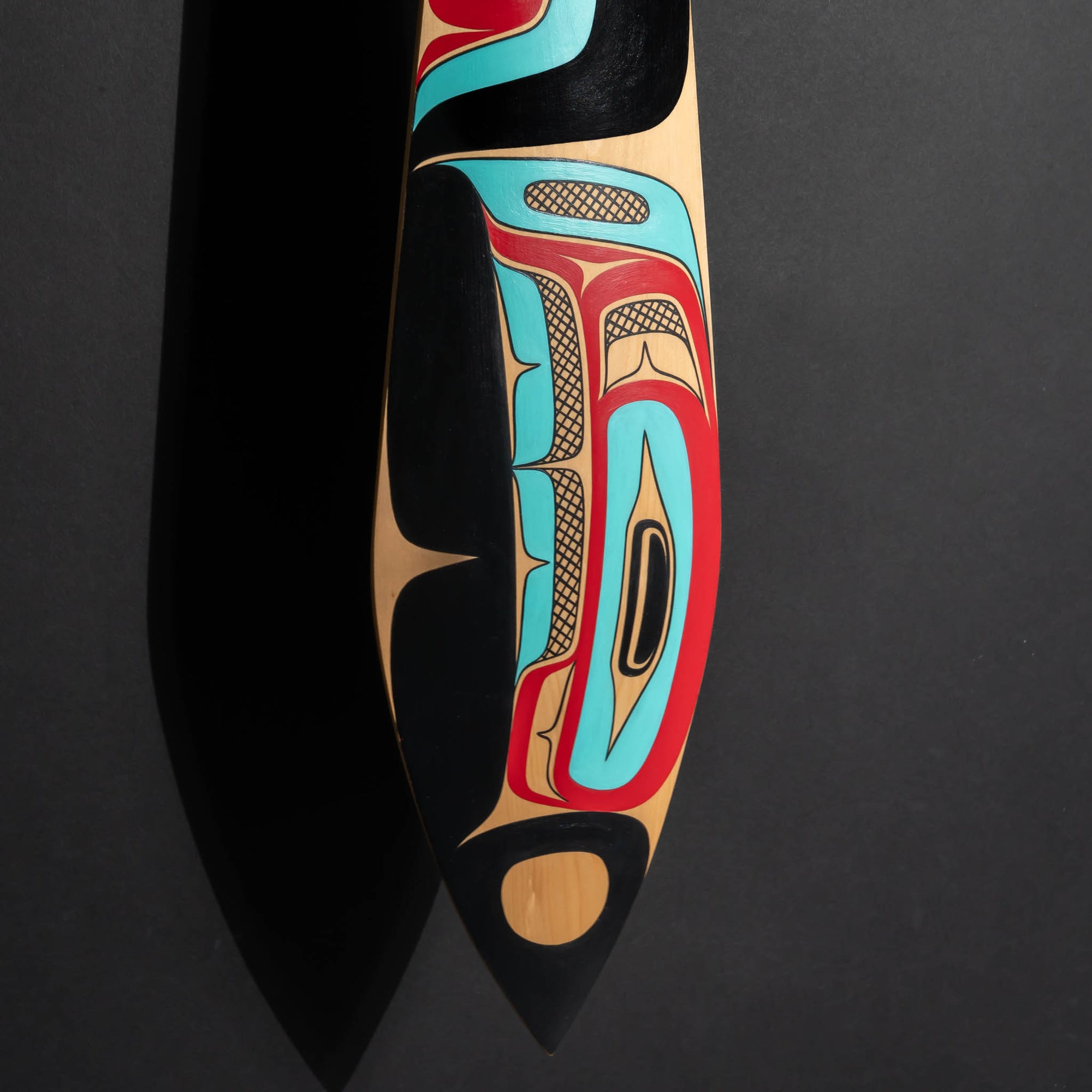 NEW: Closeup of the bottom tip of a painted paddle with black, red and blue paint with a shadow on the left and a dark background.