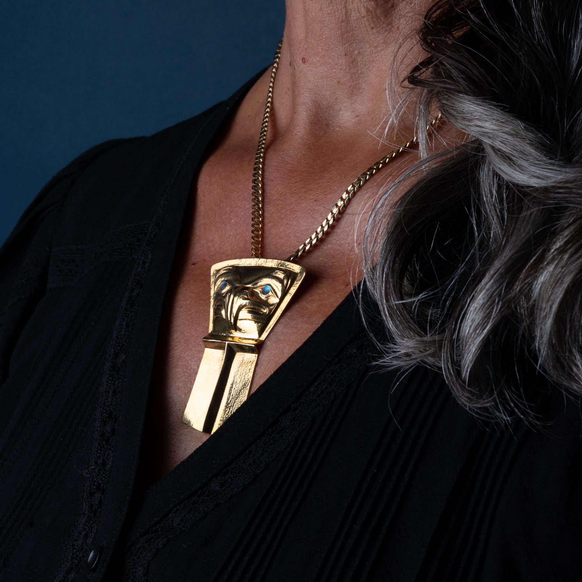 A model facing right with a black shirt and grey hair wearing a gold pendant in the shape of a shield