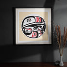 Framed artwork with a stylized formline  raven design on a beige background, placed on a dark surface.