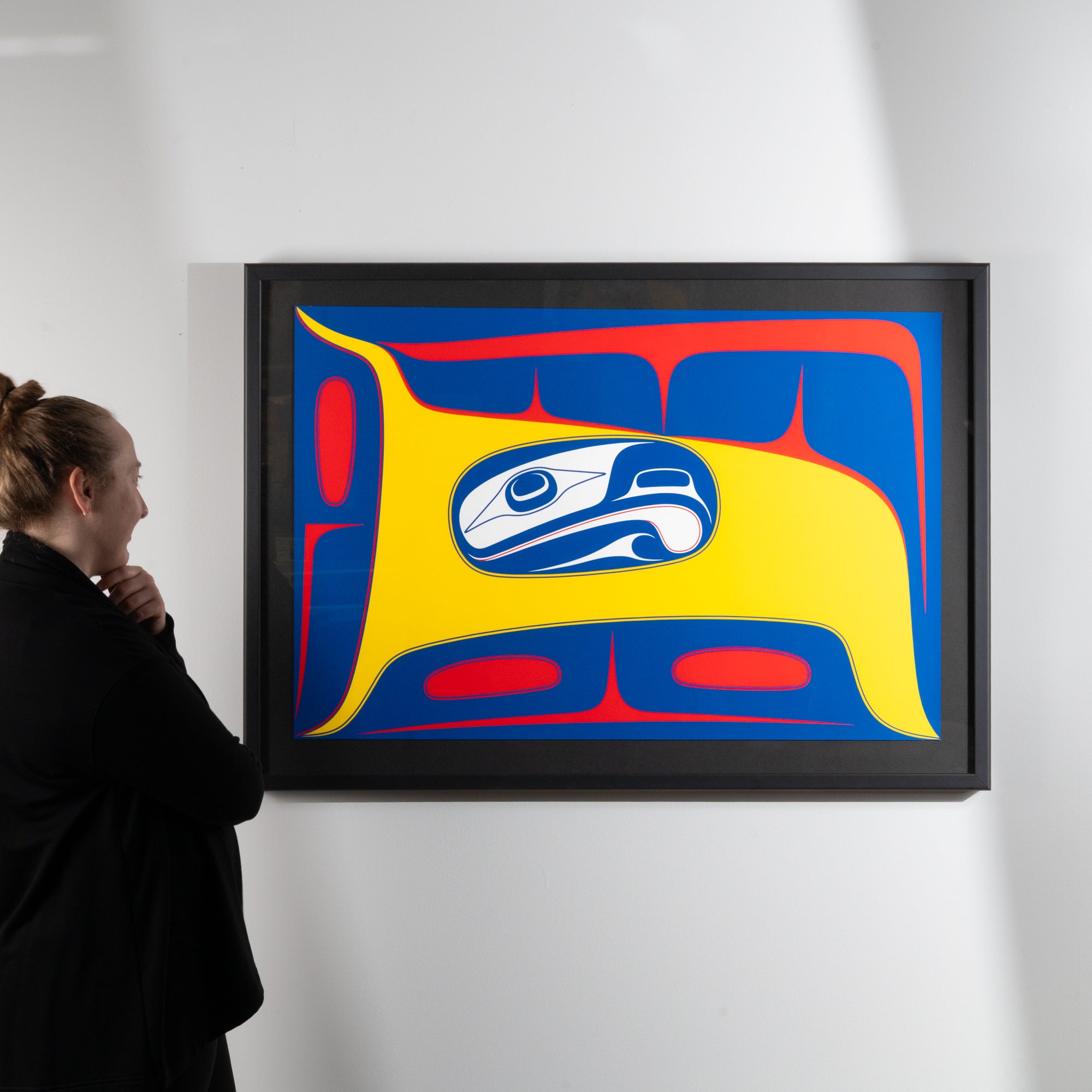 Person looking at a framed abstract artwork with a prominent blue and yellow  stylized formline design featuring a blue and white abstract eagle figure with yellow and red background by renowned haida artist Robert Davidsonon a white wall.