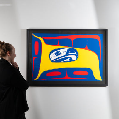 Person looking at a framed abstract artwork with a prominent blue and yellow  stylized formline design featuring a blue and white abstract eagle figure with yellow and red background by renowned haida artist Robert Davidsonon a white wall.