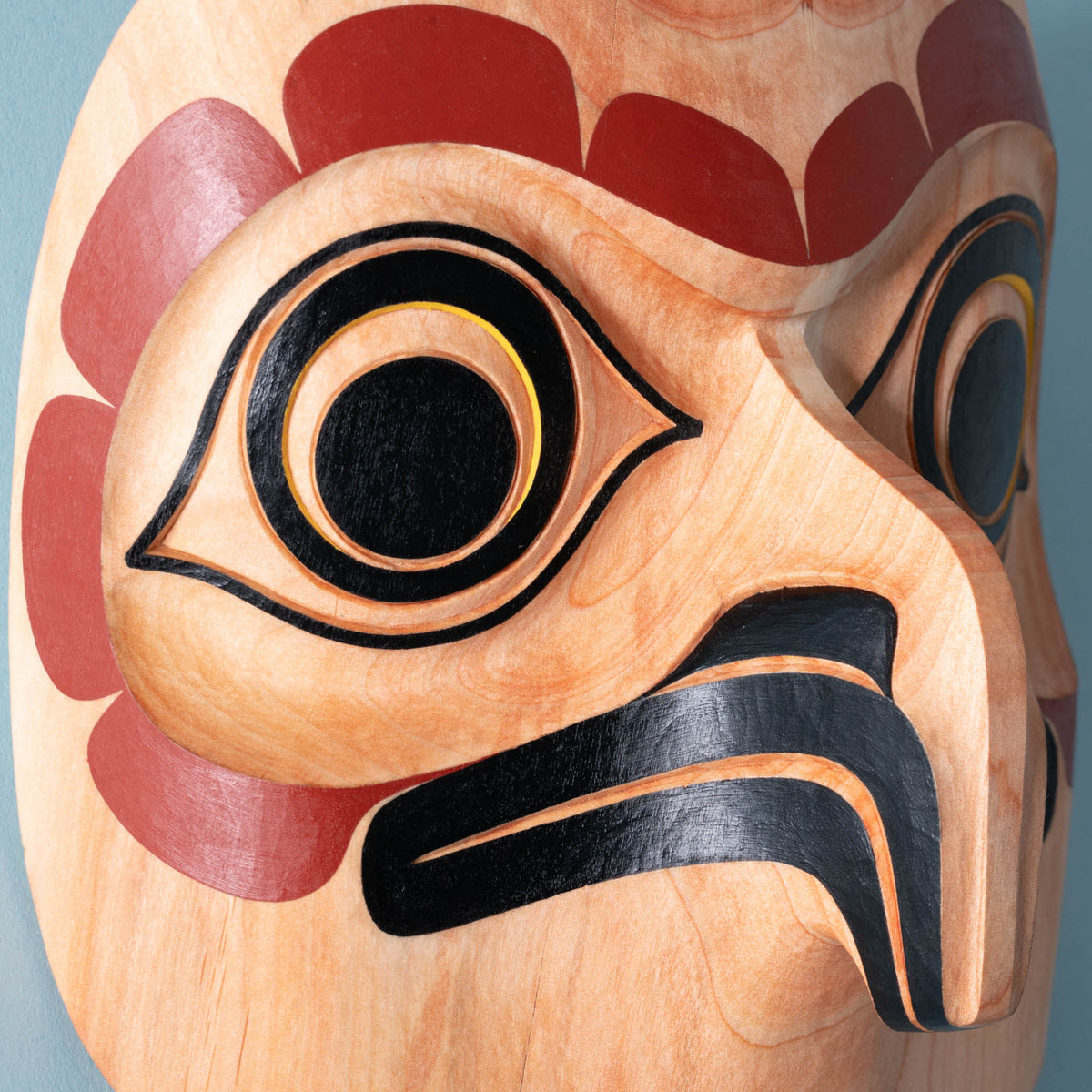 close up of eye and beak of wooden mask with intricate carvings on a light blue background.