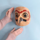 Wooden mask with owl design held by a person against a light blue. background
