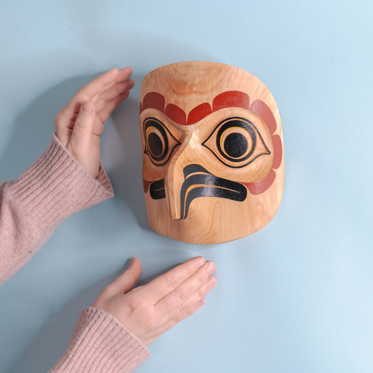 Wooden mask with owl design held by a person against a light blue. background