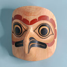 light wooden mask of an owl with red, black, and yellow paint on a blue background, showing right side of the mask.