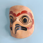 Light wooden owl mask with red, black, and yellow paint on a light blue background showing left side beak and eye.