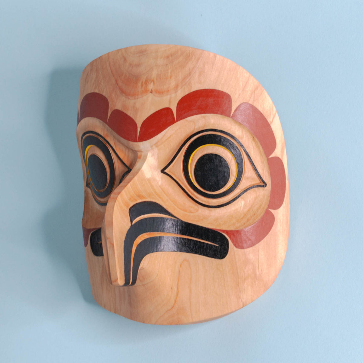 Light wooden owl mask with red, black, and yellow paint on a light blue background showing left side beak and eye.