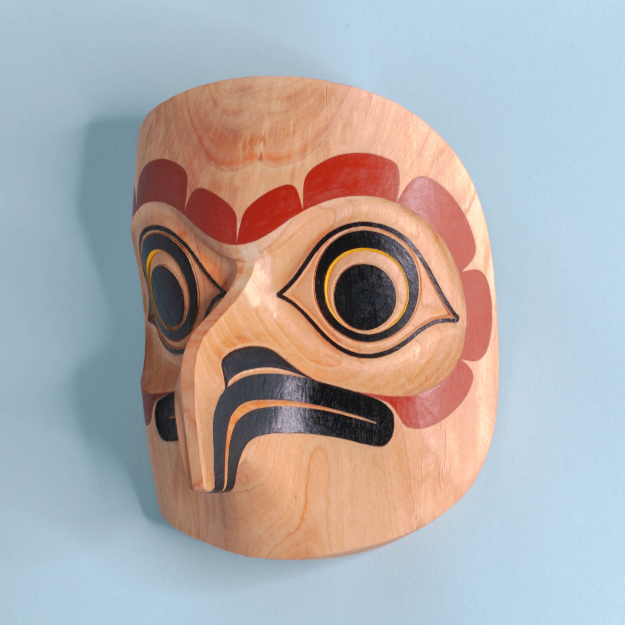 Light wooden owl mask with red, black, and yellow paint on a light blue background showing left side beak and eye.