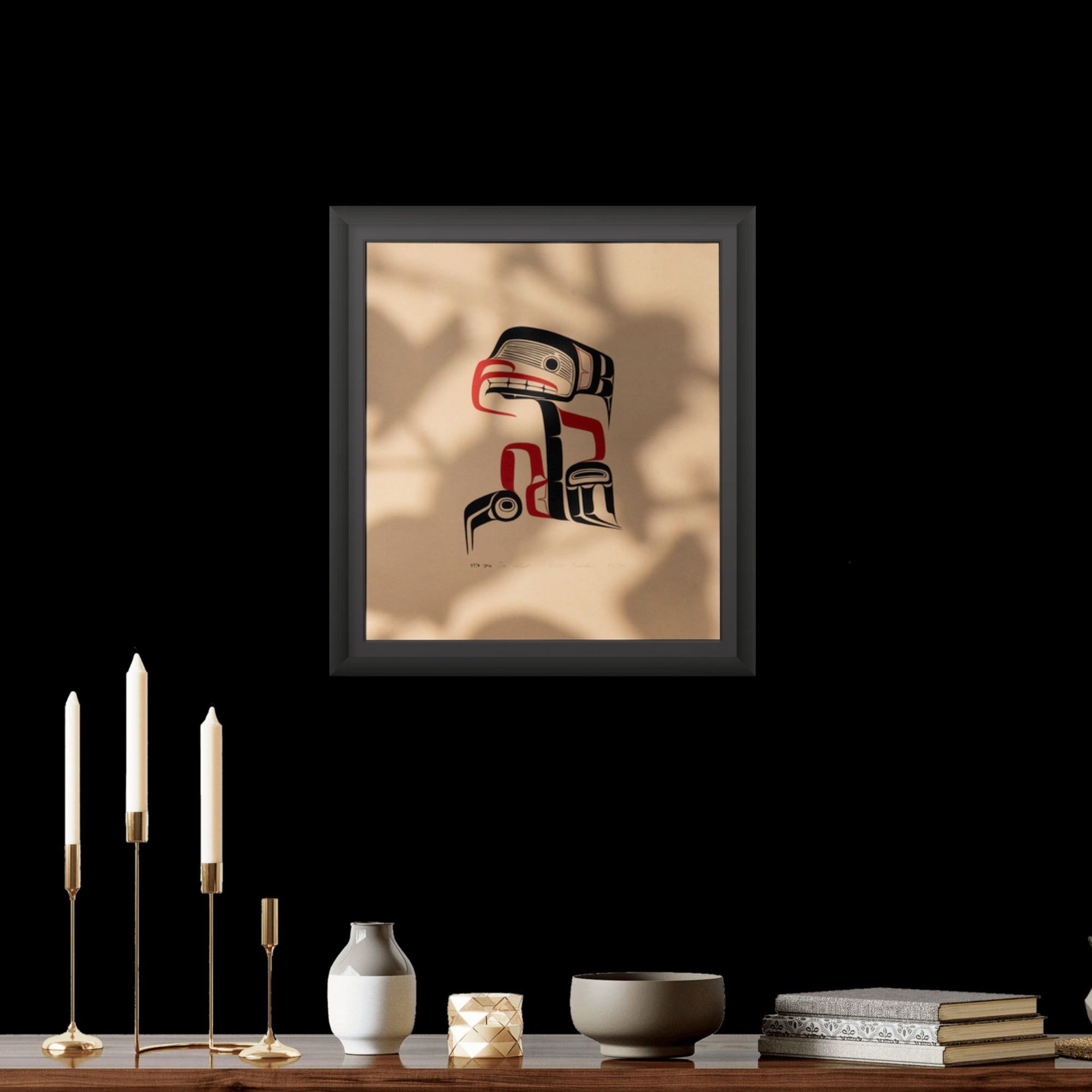 Framed artwork  depiction of a stylized Haida Sea Ghost with red and black Formline patterns on a beige background on a wall with candles and books on a table below.
