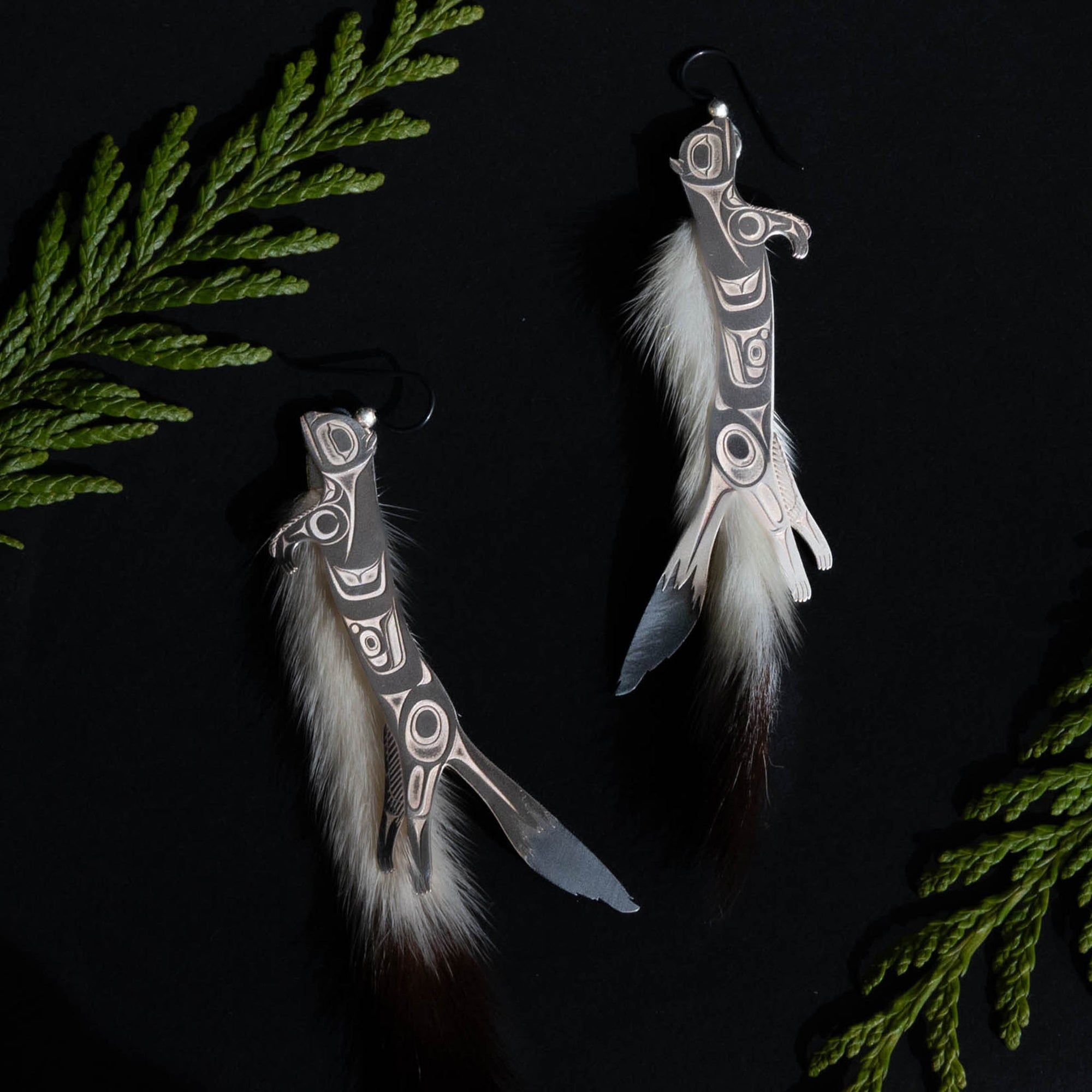 Silver ermine earrings on a black background with green cedar branches above and below.