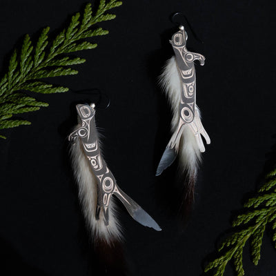Silver ermine earrings on a black background with green cedar branches above and below.