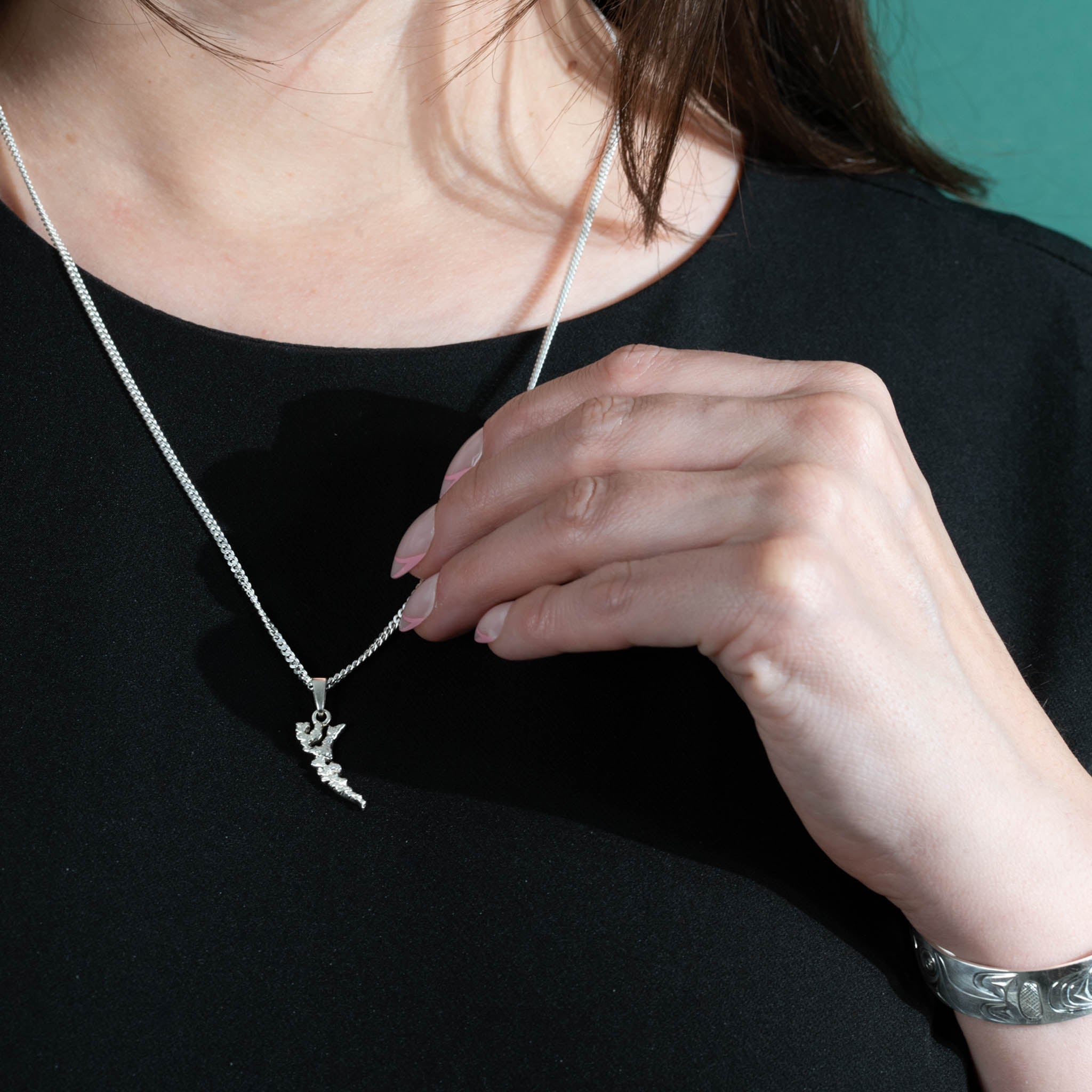 Woman wearing a silver chain and island shaped pendant with her left hand touching the chain. 