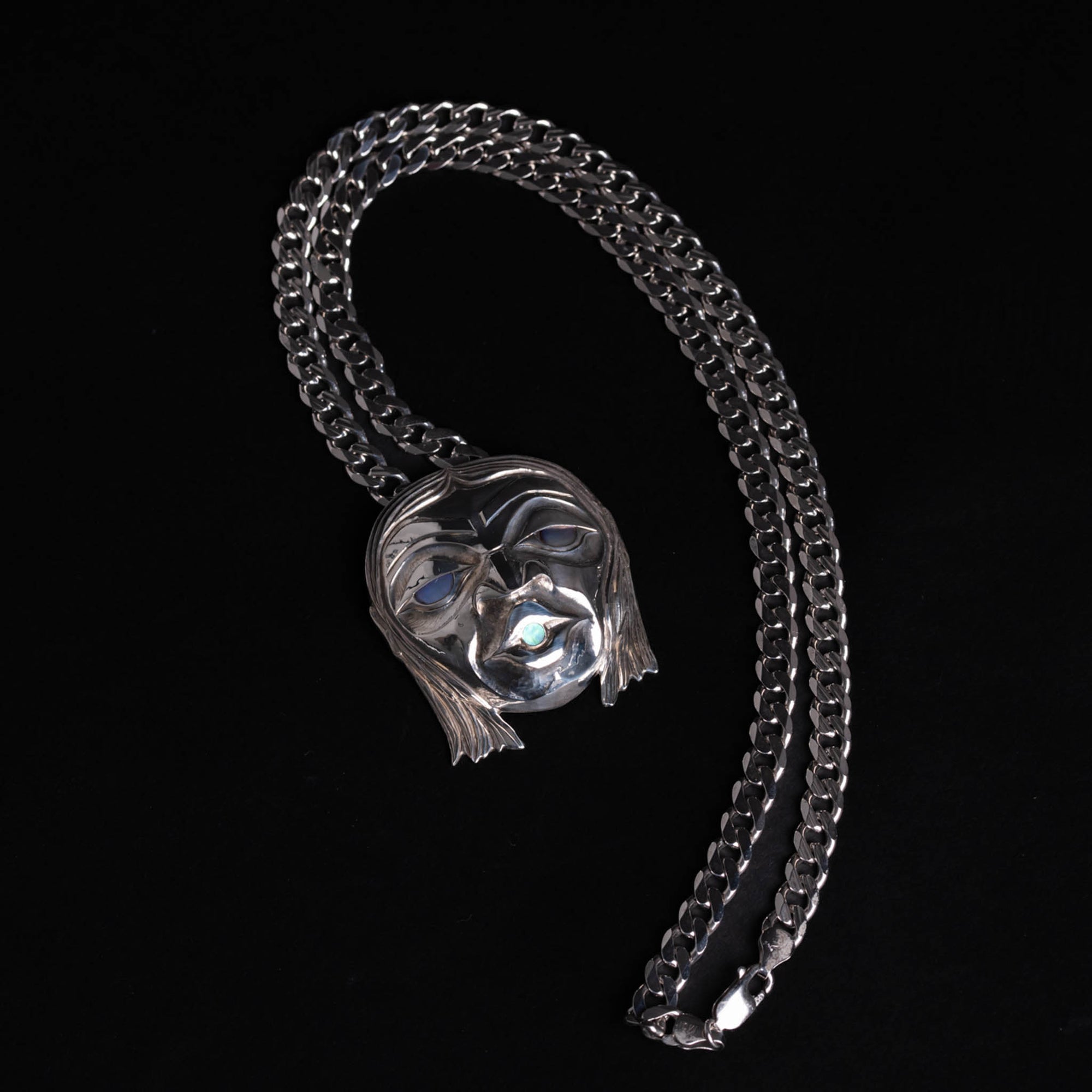 Silver mask pendant with glowing blue in the eyes and mouth with a silver chain on a black background.
