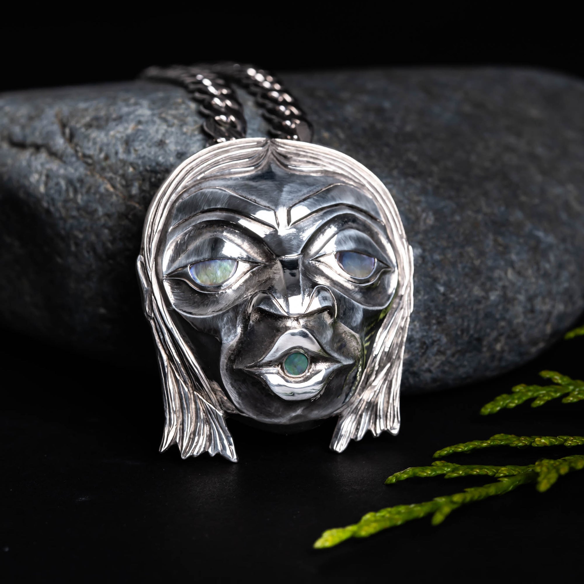 Sterling Silver Haida 'Wild Man' Repoussè Pendant by Jesse Brillon, set against a rock, with a cedar sprig, on a dark background.