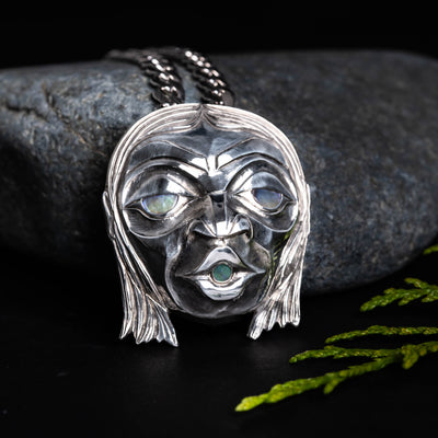 Sterling Silver Haida 'Wild Man' Repoussè Pendant by Jesse Brillon, set against a rock, with a cedar sprig, on a dark background.