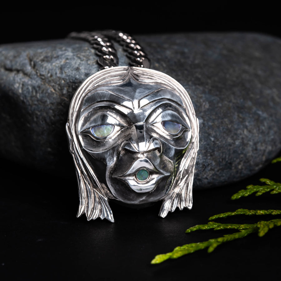 Sterling Silver Haida 'Wild Man' Repoussè Pendant by Jesse Brillon, set against a rock, with a cedar sprig, on a dark background. 