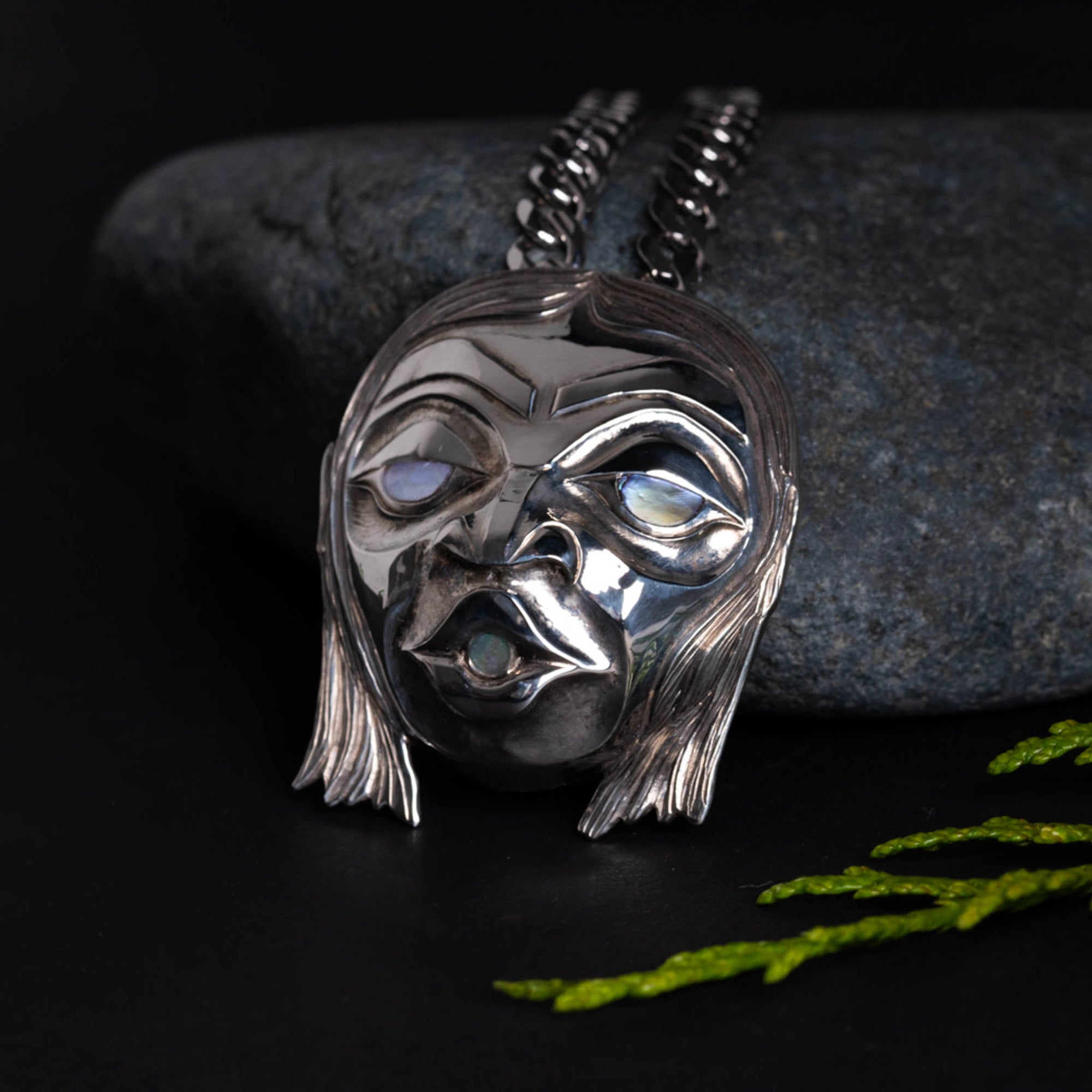 Silver mask like pendant with glowing eyes, leaning against a grey rock on a dark backgroung.