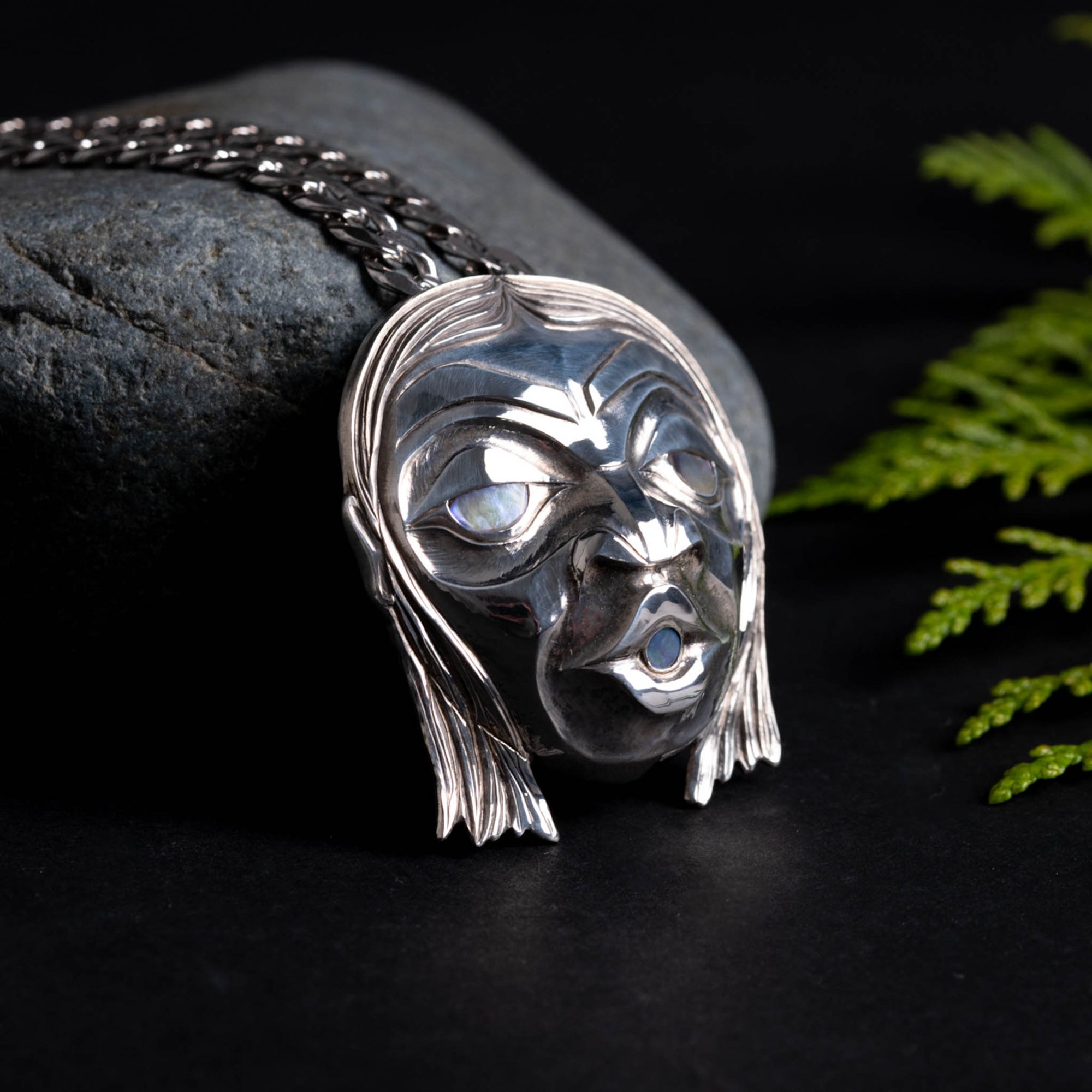 Silver pendant of a face with long hair and glowing eyes, with a silver chain. on a dark surface with greenery.