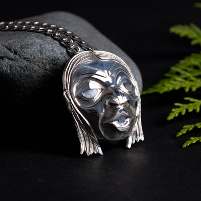 Silver pendant of a face with long hair and glowing eyes, with a silver chain. on a dark surface with greenery.