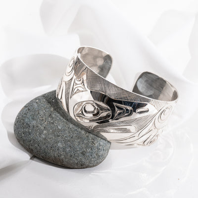 Front of Sterling Silver Haida Raven bracelet with intricate formline designs hand carved by James Sayer Haida Jeweller on a gray stone against a white background