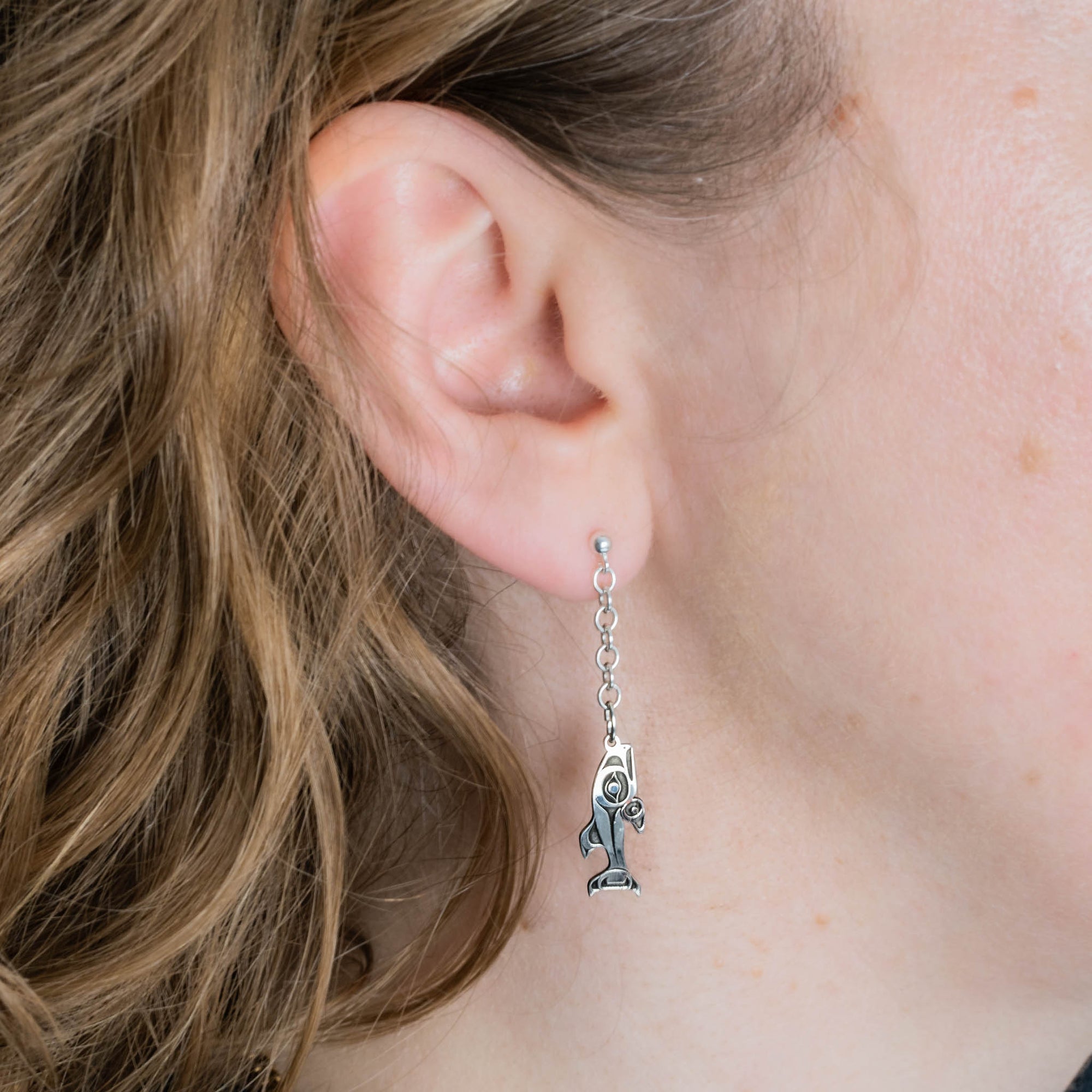 Close-up of an ear wearing a silver earring with a fish charm and chain.