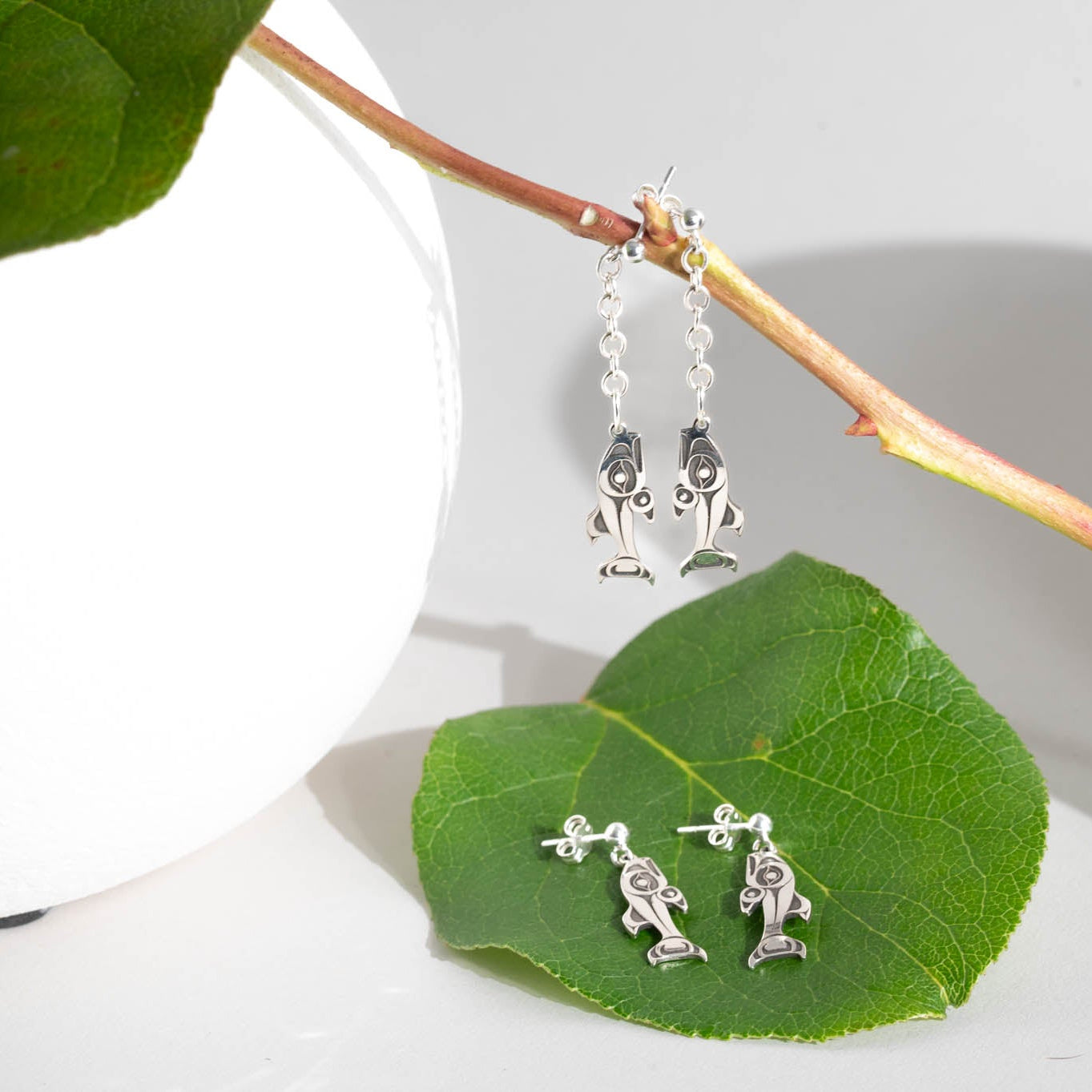 Silver salmon-shaped earrings on a green leaf with a white vase and branch in the background.