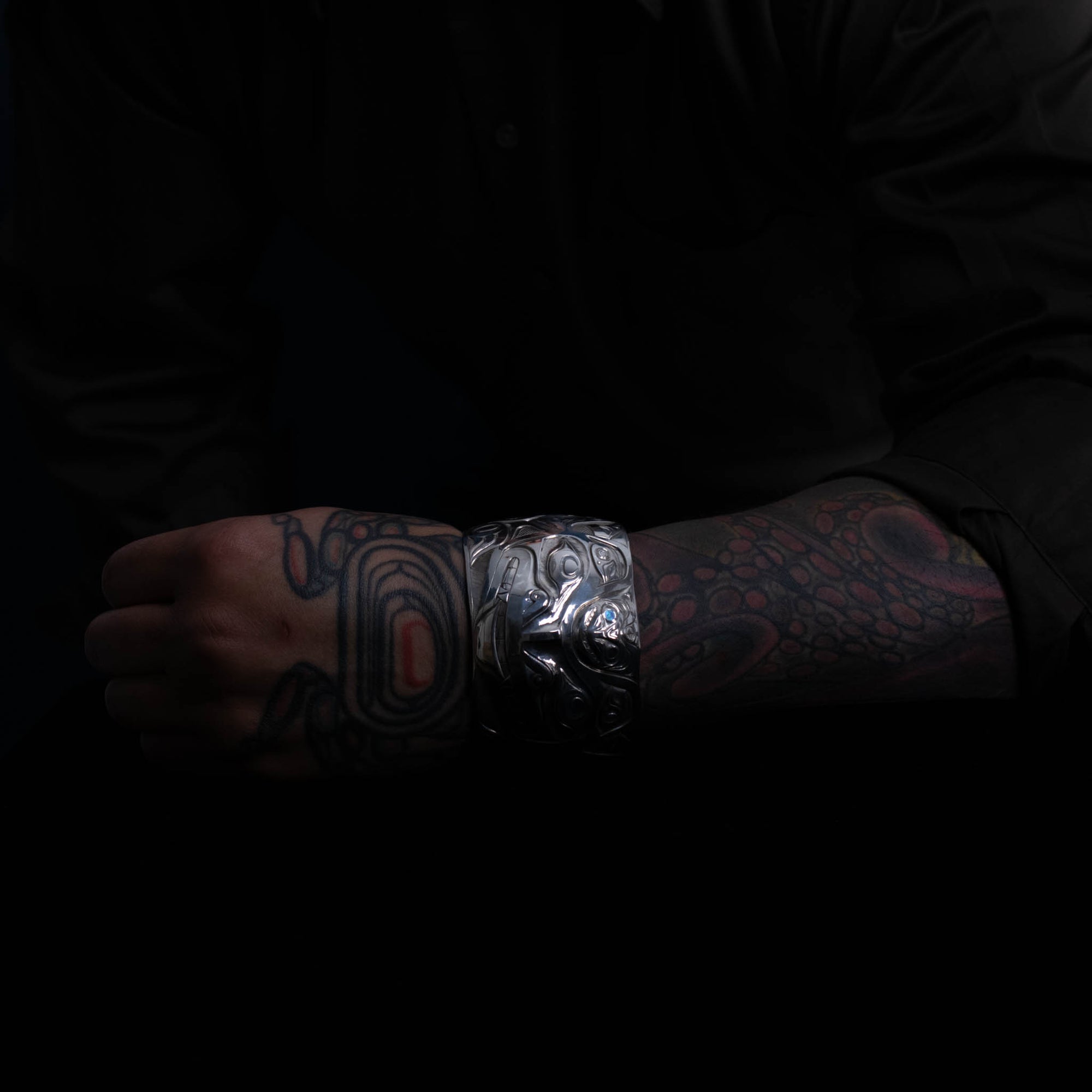 Person's arm with tattoos wearing a metallic bracelet against a dark background