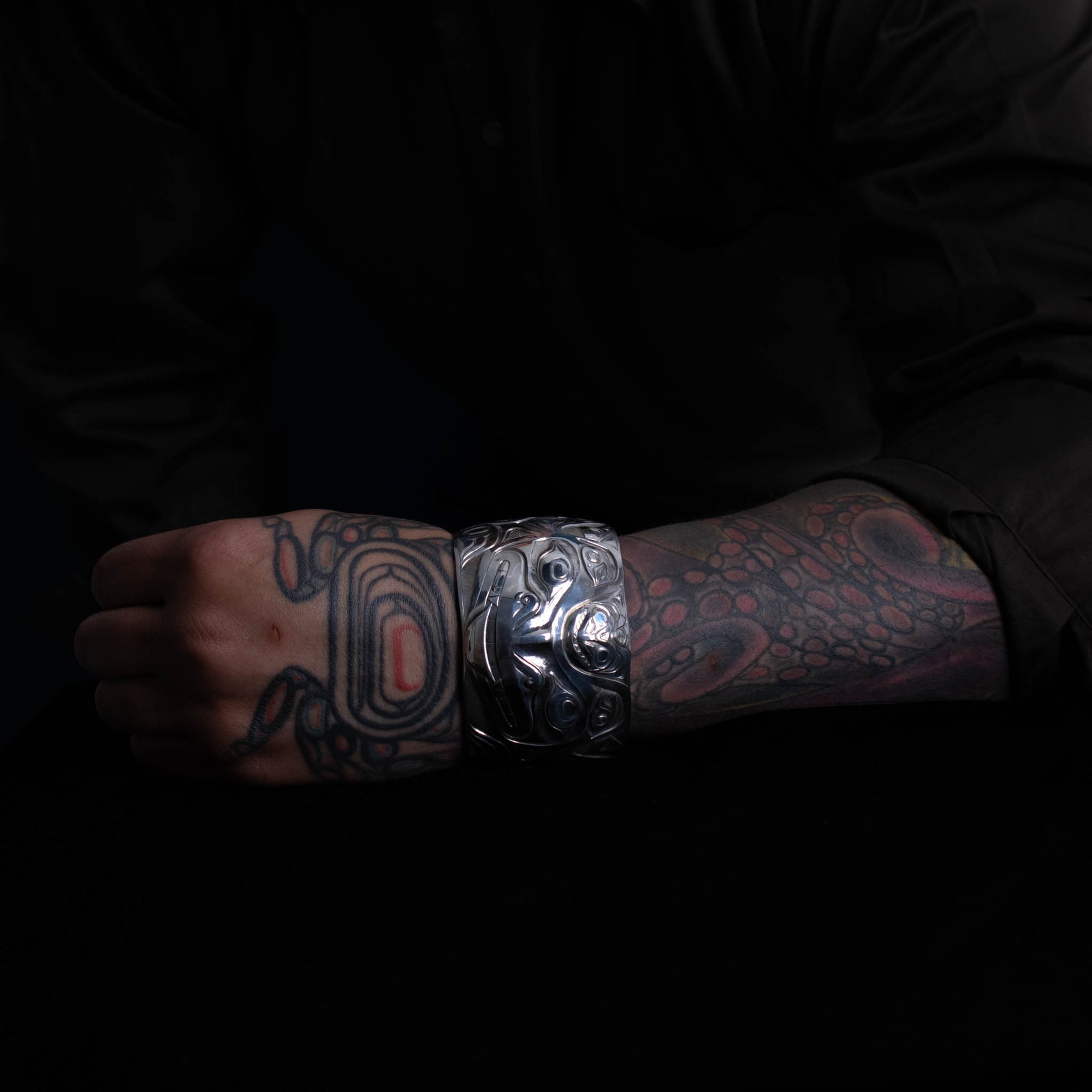 Person with tattoos wearing a silver bracelet by Jesse Brillon with intricate northwest coast design on a dark background