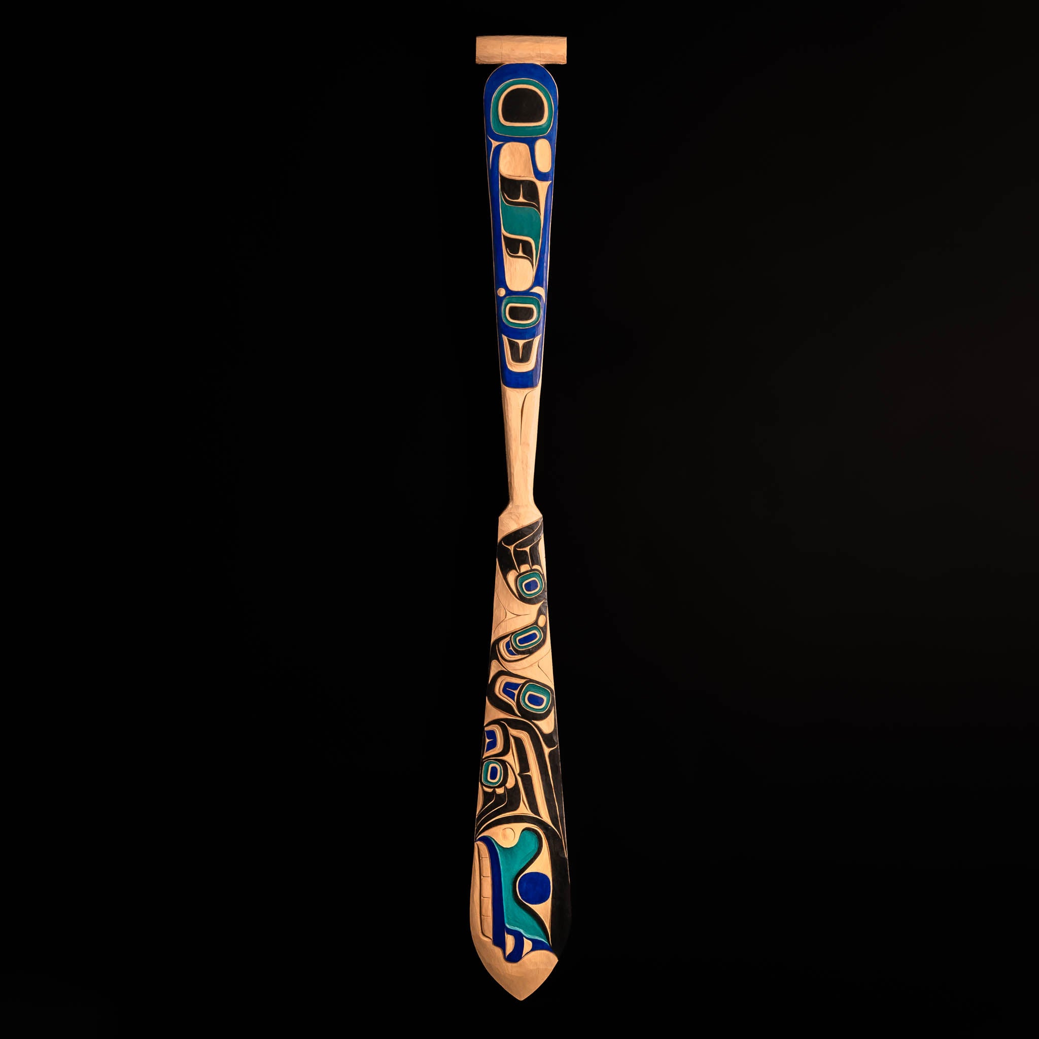 Decorative wooden paddle with intricate blue and black Haida patterns on a black background.